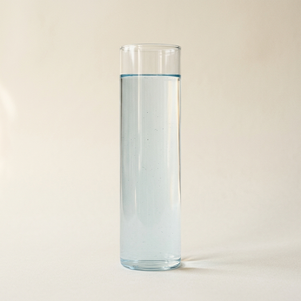 Tall glass cylinder of tap water with a faint blue chlorinated tint, on a cream background